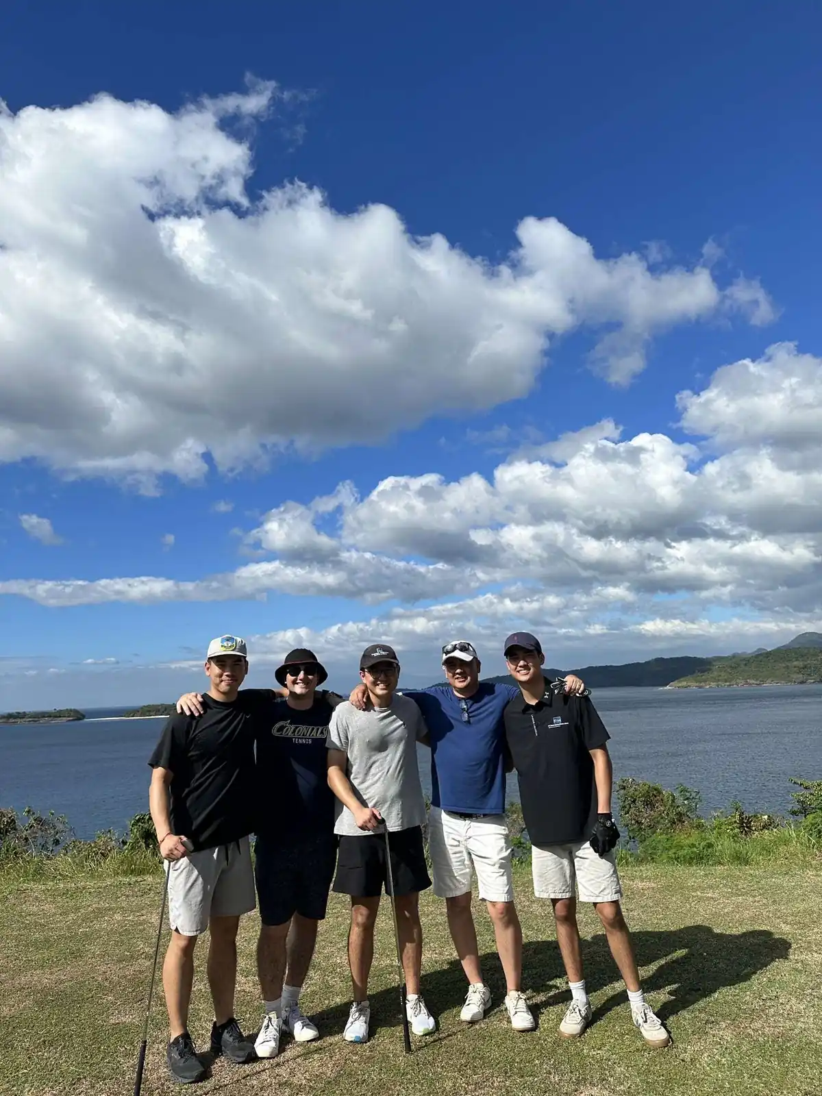 Five people standing together on a golf course beside the water